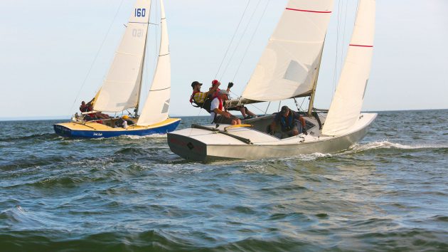 Two sailing boats floating away on relatively calm seas. In the boat closest to the camera, are leaning over the side in a racing position. The Yachting World Diamond is streaming through the water.