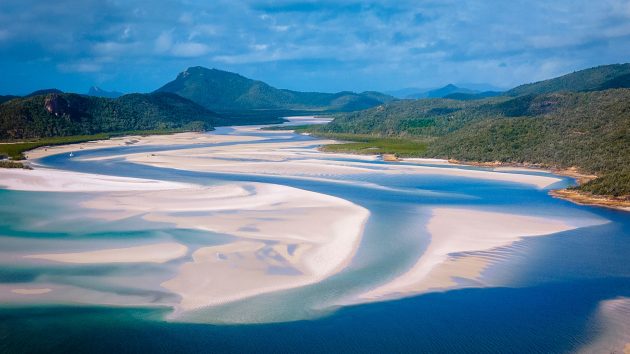 Aerial shot of a sand bank with rivers running through it, mountains in the background, and blue sky.